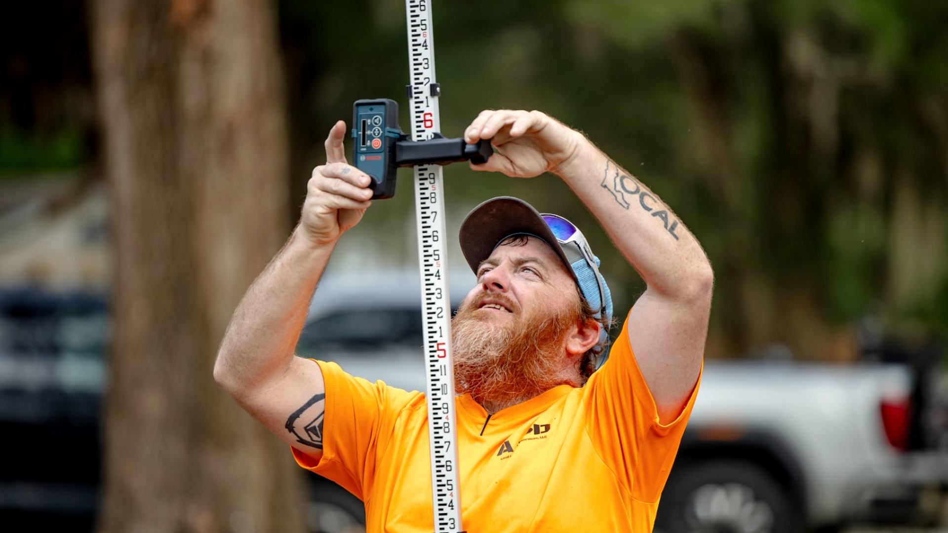Dusty Tyer holding a laser receiver against a grade rod on a job site under live oaks in Central Florida